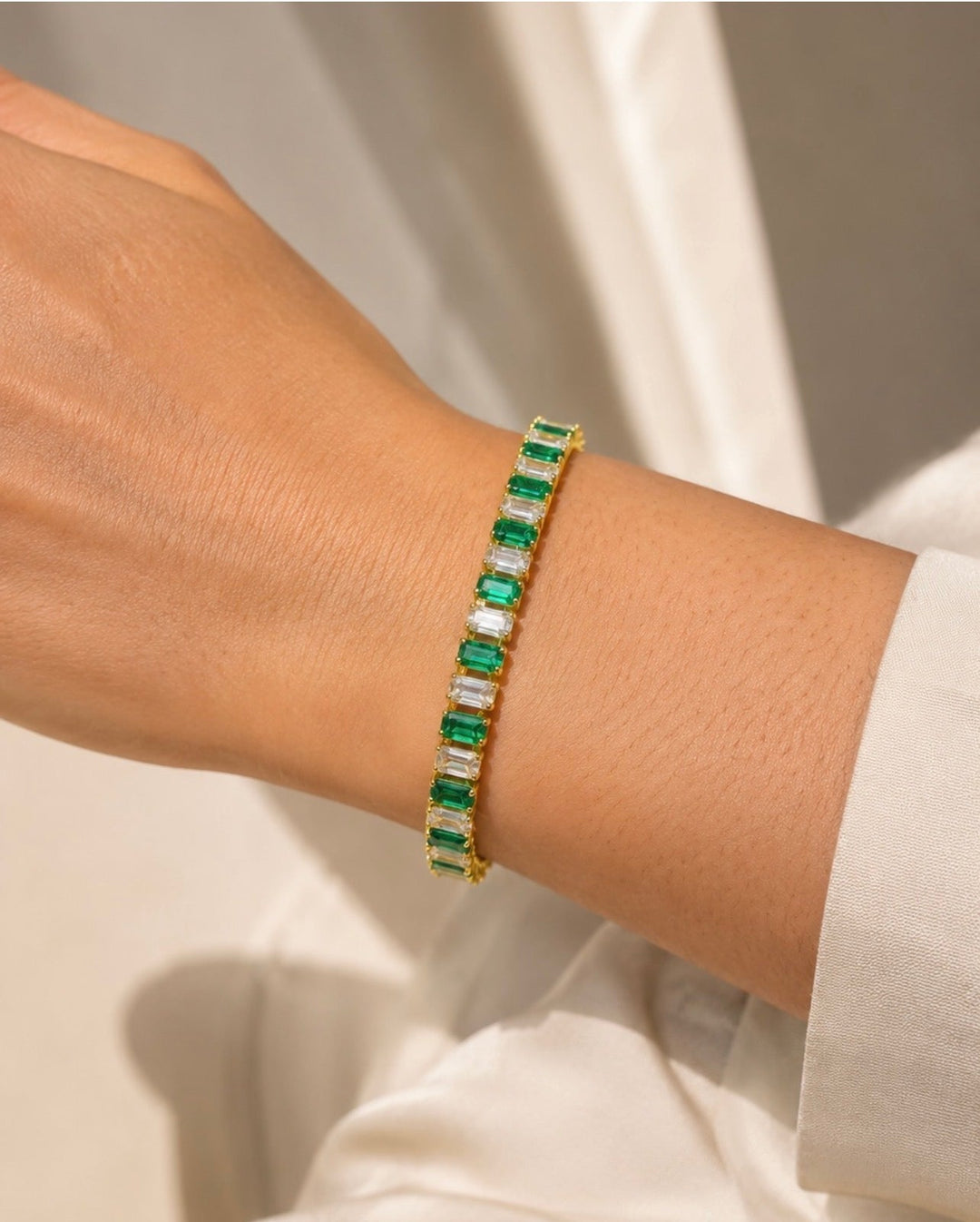 Bracelet Ahmès doré, alternant pierres rectangulaires vertes et transparentes, porté au poignet près d’une manche blanche.