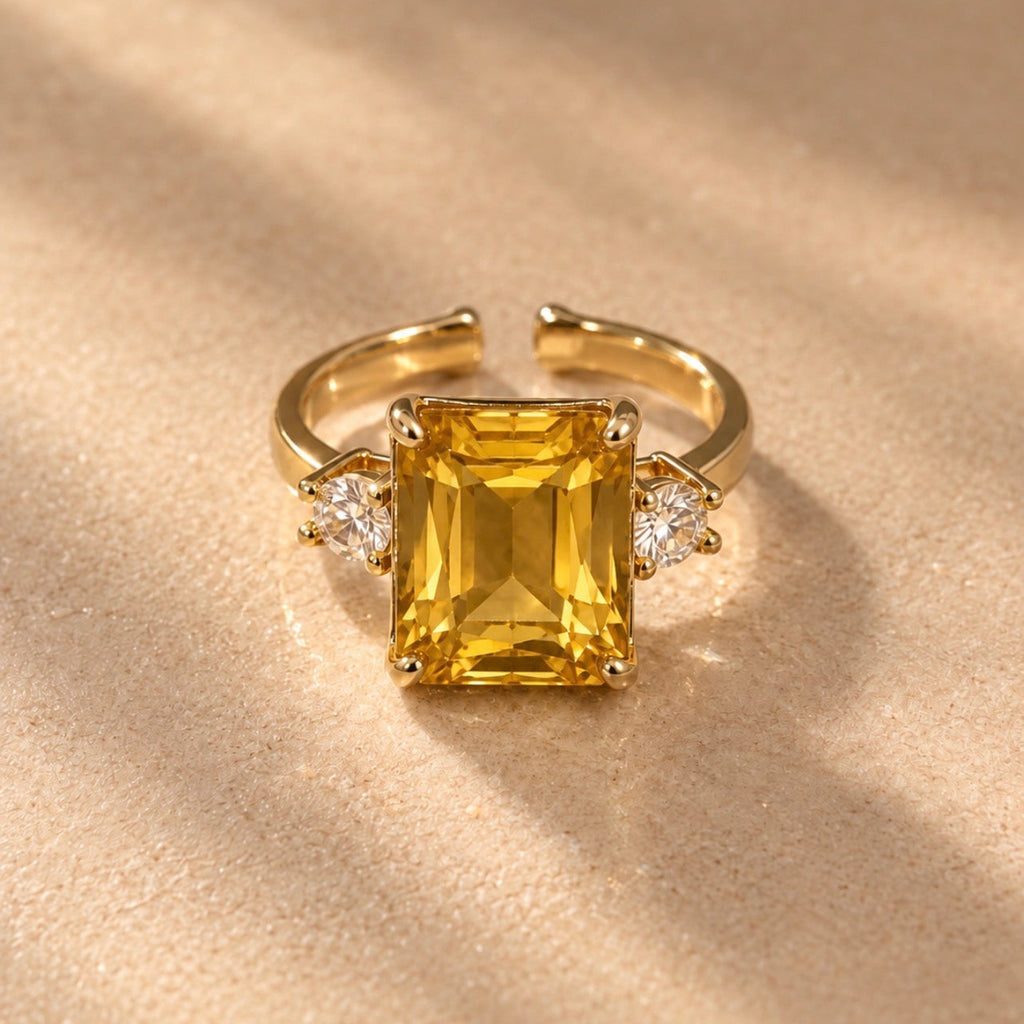 Bague Iset dorée à nano-saphir jaune rectangulaire et cristaux latéraux, posée sur un tissu beige