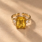 Bague Iset dorée à nano-saphir jaune rectangulaire et cristaux latéraux, posée sur un tissu beige