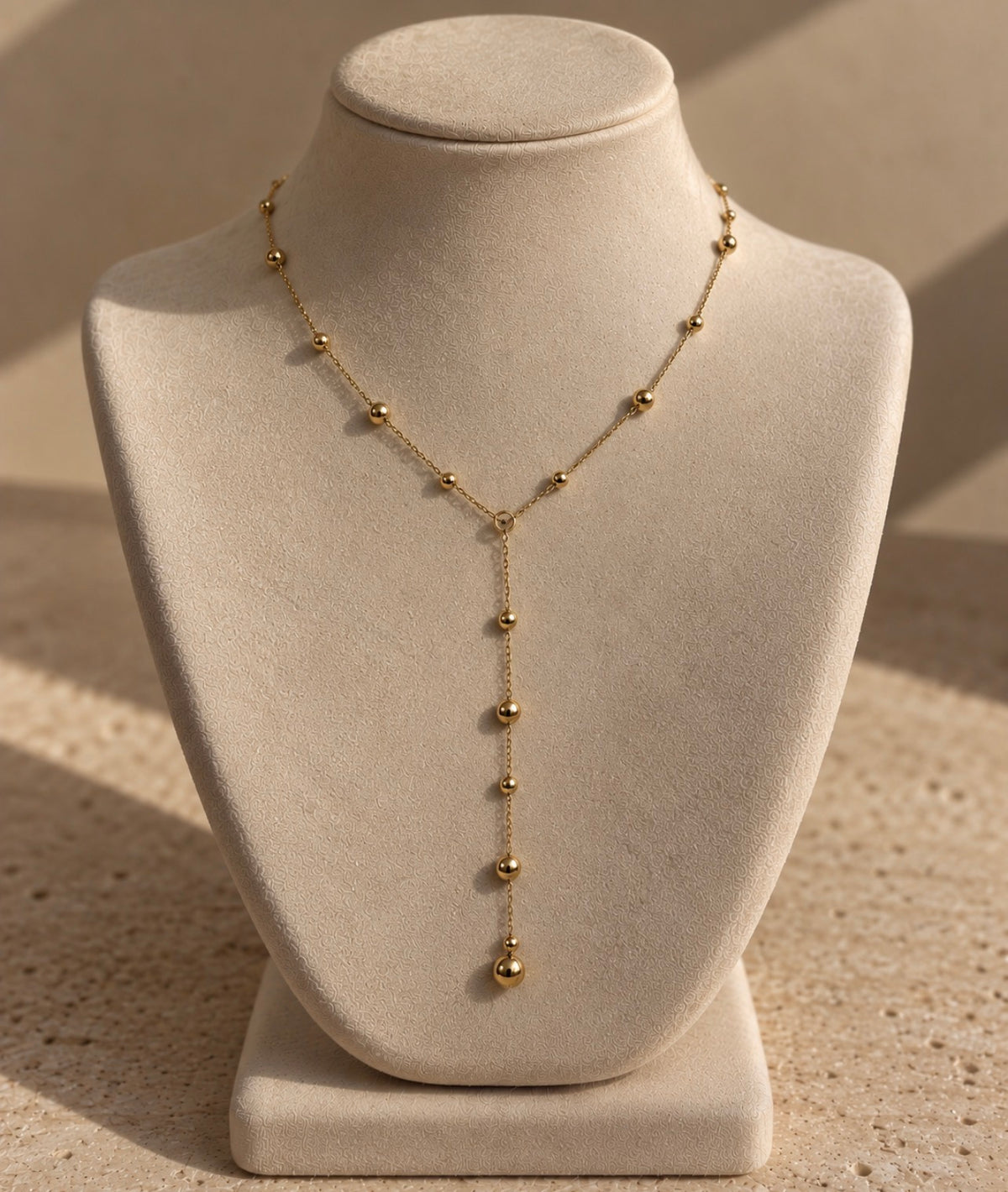 Collier Néfertari en laiton doré à l’or 18 carats, chaîne en Y ornée de boules dorées, présenté sur un buste crème.