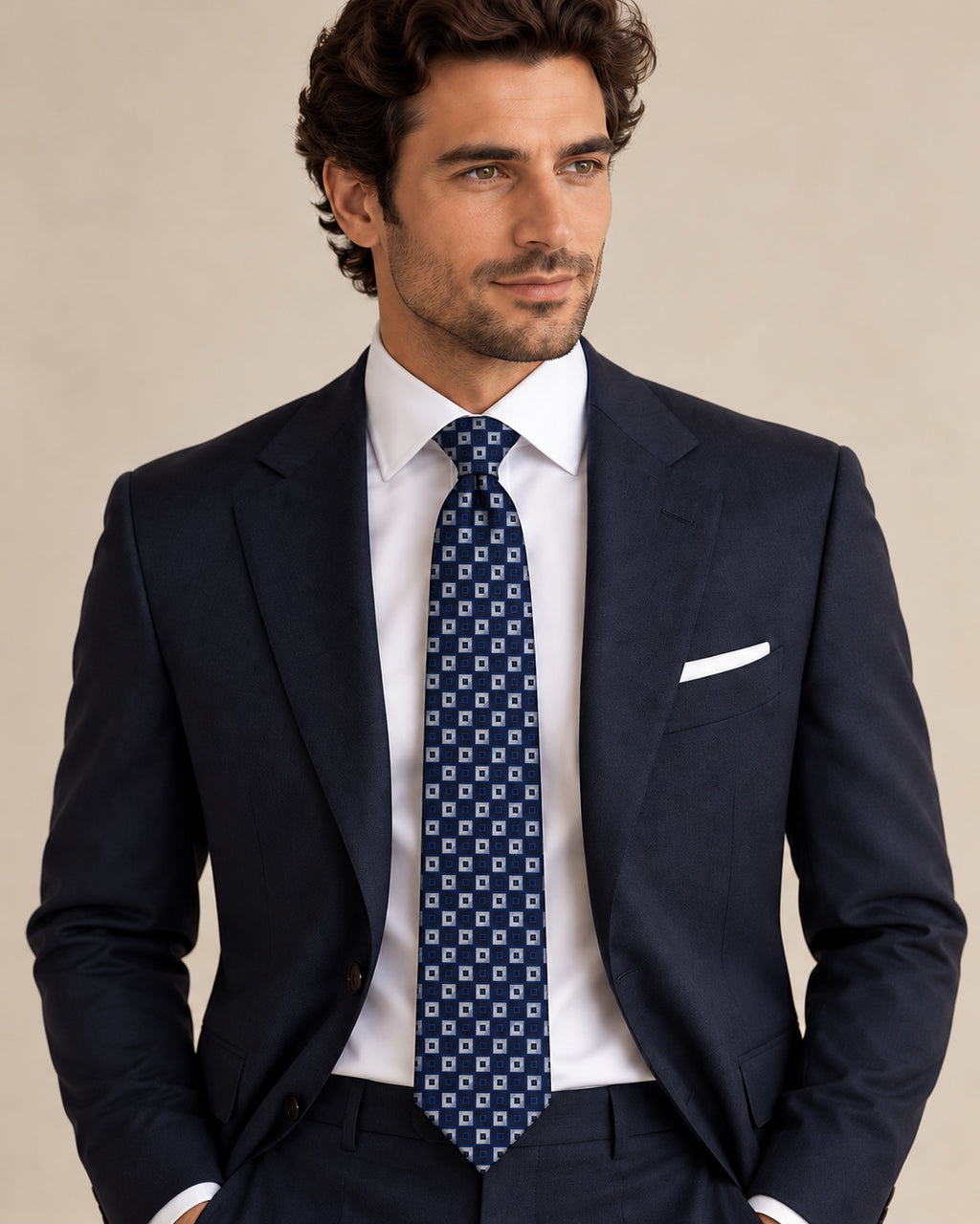 Cravate Gramont à carrés bleus et gris argenté, portée avec un costume bleu marine, une chemise et une pochette blanches.