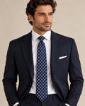 Cravate Gramont à carrés bleus et gris argenté, portée avec un costume bleu marine, une chemise et une pochette blanches.