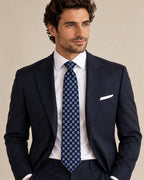 Cravate Gramont à carrés bleus et gris argenté, portée avec un costume bleu marine, une chemise et une pochette blanches.