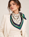 Foulard Rue de l’Éperon en soie ivoire à mors équestres noirs et bordure vert émeraude et bleu marine, porté sur un tailleur crème.